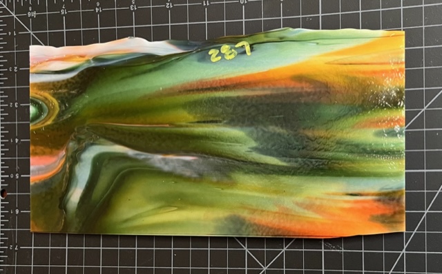 A rectangle of sheet glass on a black cutting surface. The glass contains brightly contrasting streaks of color in shades of green, orange, white, and yellow.